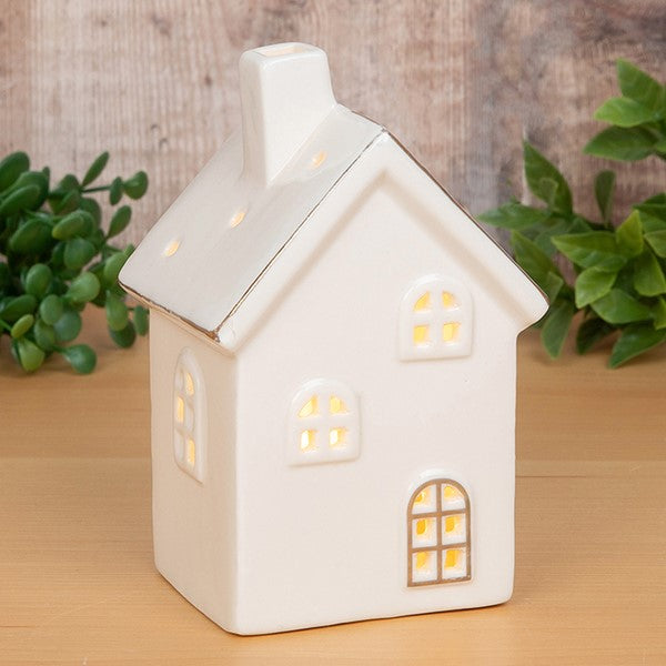 Cosy Lane LED light-up house ornament small, glowing with warm golden light through cut-out windows, gold trim detail on roofline and arched door, styled on a wooden shelf with greenery