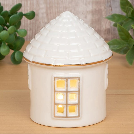 Cosy Lane LED light-up round cottage small, glowing with warm golden light through cut-out window, textured conical roof with gold trim, styled on a wooden shelf with greenery