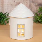 Cosy Lane LED light-up round cottage medium, glowing with warm golden light through a cut-out window, textured conical roof with gold trim, styled on a wooden shelf with greenery