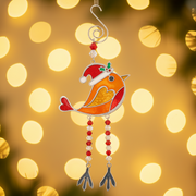 A stained glass style Christmas robin decoration in orange and red with a red Santa hat and holly, dangly red and clear bead legs, and a silver spiral hanging hook, displayed with warm golden fairy light bokeh