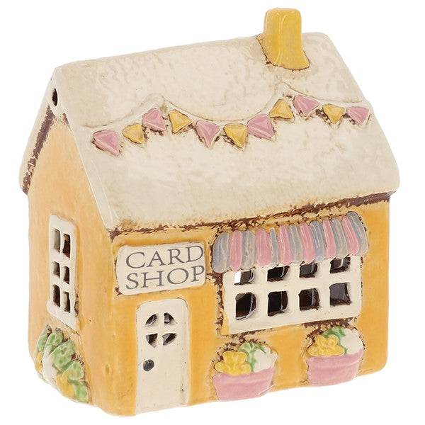 Village Pottery Card Shop ceramic tealight holder with yellow exterior, striped awning, chimney pot, flags and plants by the door, front view on white background