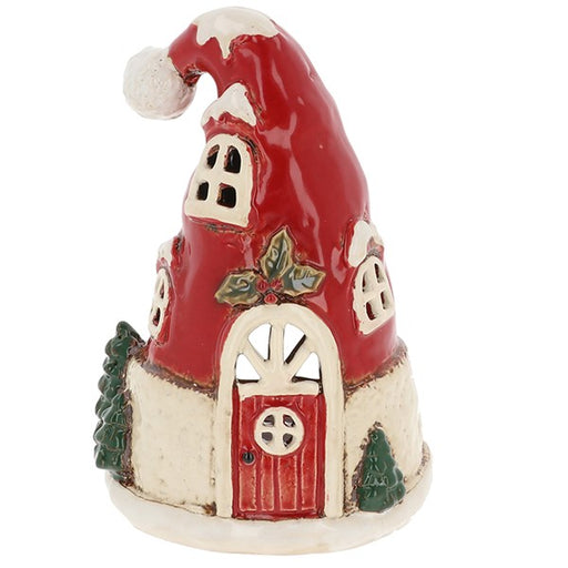 Village Pottery Christmas Santa hat house tealight, red drooping hat roof with white pompom and brim trim, holly and berry sprig above door, round fanlight window, red panelled door and Christmas trees at base, white background