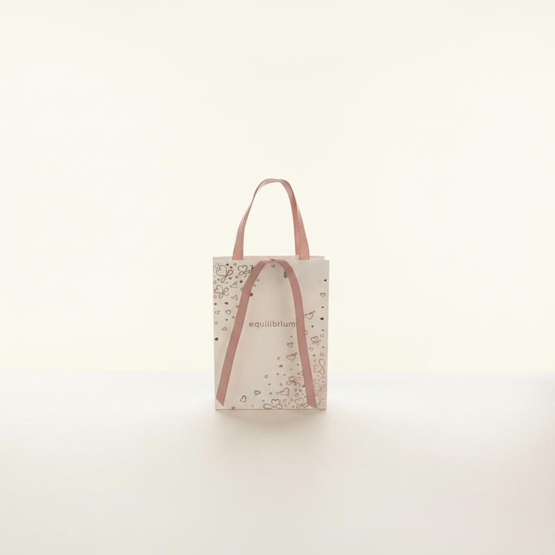 A white paper gift bag printed with a rose gold foil heart pattern with "equilibrium" text and dusky pink ribbon handles