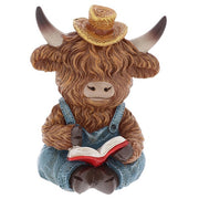 Highland cow reading figurine sitting upright in denim dungarees and straw hat, reading a red-covered book, shaggy fur and curved horns