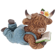 Highland cow reading figurine lying on her front in denim dungarees with flower hair accessories, reading a green-covered book, shaggy fur and curved horns