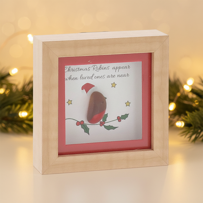 A small square natural wood box framed pebble art picture with a pebble robin wearing a red Santa hat perched on an illustrated holly branch with gold stars, and the sentiment "Christmas Robins appear when loved ones are near", displayed with warm golden fairy light bokeh