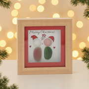 A small square natural wood box framed pebble art picture with two pebble figures wearing red Santa hats standing beneath a sprig of mistletoe with red hearts and the sentiment "Merry Christmas", displayed with warm golden fairy light bokeh