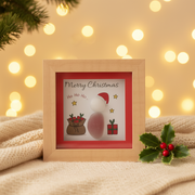 A small square natural wood box framed pebble art picture with a pebble Santa figure, illustrated gift sack, wrapped present and gold stars, with the sentiments "Merry Christmas" and "Ho Ho Ho...", displayed with warm golden fairy light bokeh