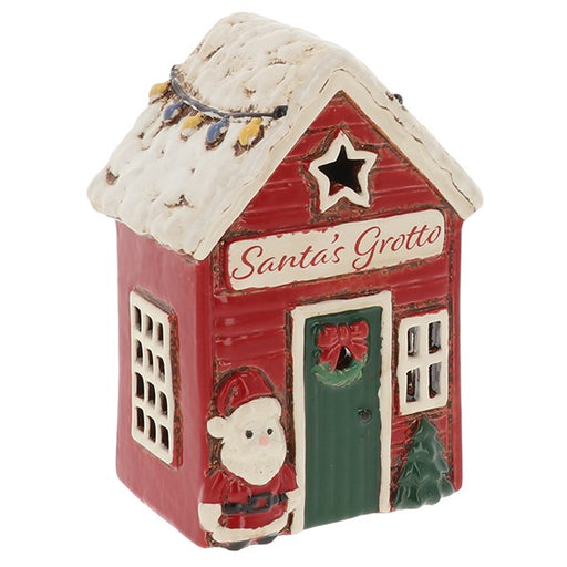 Village Pottery Santa's Grotto tealight holder — red cottage with snow-dusted roof, colourful fairy lights, star cut-out in the gable, "Santa's Grotto" sign, green door with wreath and bow, Father Christmas figure to one side and Christmas tree to the other