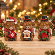 Three Merry Christmas Coos mini highland cow figurines displayed together, holding a bauble, a gingerbread house and a wrapped present, with warm golden bokeh fairy lights background