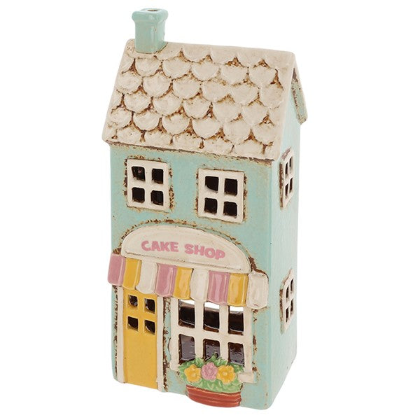 Angled view of Village Pottery cake shop tea light holder showing duck egg blue chimney, scalloped roof detail and terracotta flower pot by the door