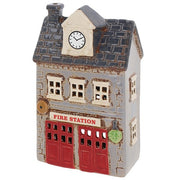 Angled view of Village Pottery fire station tea light holder showing clock tower, chimney, brass bell detail and green plant by the entrance