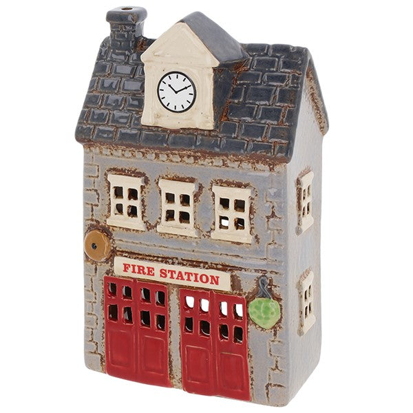 Angled view of Village Pottery fire station tea light holder showing clock tower, chimney, brass bell detail and green plant by the entrance