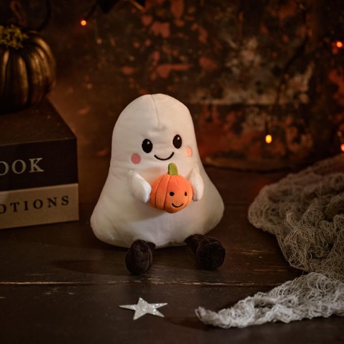 Soft white plush ghost beanie with smiling face, rosy cheeks and black legs holding a small orange pumpkin, styled on a dark Halloween-themed background with fairy lights