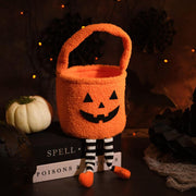 Pumpkin Halloween treat bag with jack-o'-lantern face, carry handle and black-and-white striped dangly legs with orange feet, styled on a dark Halloween-themed background with fairy lights and a white pumpkin