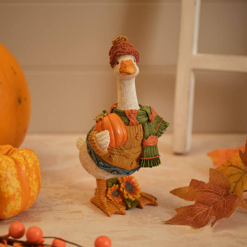 Standing duck ornament dressed in a bobble hat, green scarf and jumper, holding a pumpkin with sunflowers at her feet, autumn seasonal decoration