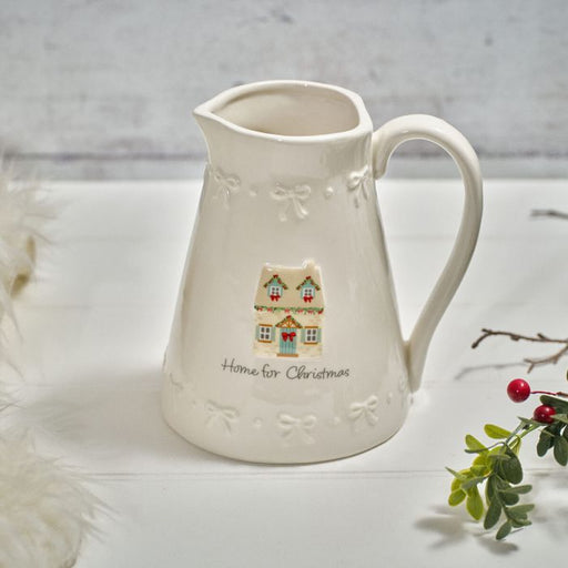 Cream stoneware Home for Christmas decorative jug with embossed bows and raised cottage illustration, styled with foliage on a white surface