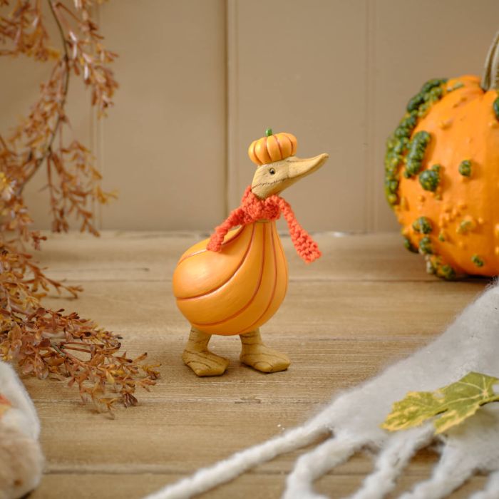 Pumpkin duck resin figurine with orange pumpkin-shaped body, pumpkin hat with green stem and red fluffy knitted scarf, styled on a wooden surface with autumn foliage and pumpkins