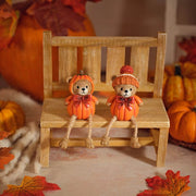 Two pumpkin bear shelf sitter ornaments seated on a wooden bench with dangly rope legs, styled with autumn pumpkins and fallen leaves