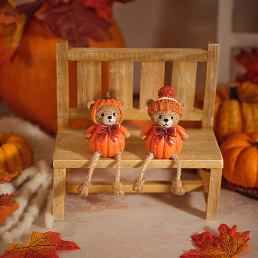 Two pumpkin bear shelf sitter ornaments seated on a wooden bench with dangly rope legs, styled with autumn pumpkins and fallen leaves