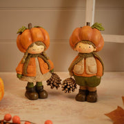 Two resin pumpkin figurines standing together — a girl with plaited hair in a textured top and lace-trim skirt, and a boy in a textured gilet and trousers, each wearing a pumpkin hat and holding a pinecone, styled on a surface with autumn leaves and berries in the background
