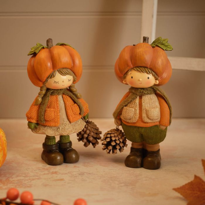 Two resin pumpkin figurines standing together — a girl with plaited hair in a textured top and lace-trim skirt, and a boy in a textured gilet and trousers, each wearing a pumpkin hat and holding a pinecone, styled on a surface with autumn leaves and berries in the background
