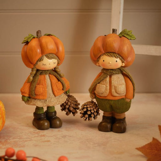 Two resin pumpkin figurines standing together — a girl with plaited hair in a textured top and lace-trim skirt, and a boy in a textured gilet and trousers, each wearing a pumpkin hat and holding a pinecone, styled on a surface with autumn leaves and berries in the background