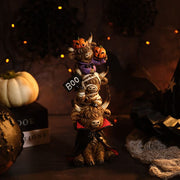 A resin triple stacking highland cow Halloween figurine with three characters — a vampire cow at the bottom in a black and red cape, a mummy cow in the middle holding a BOO sign, and a pumpkin cow at the top in a purple outfit with two jack-o'-lanterns on its horns, styled on a dark surface with Halloween decorations and warm fairy lights in the background