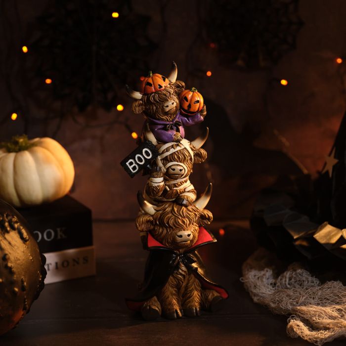 A resin triple stacking highland cow Halloween figurine with three characters — a vampire cow at the bottom in a black and red cape, a mummy cow in the middle holding a BOO sign, and a pumpkin cow at the top in a purple outfit with two jack-o'-lanterns on its horns, styled on a dark surface with Halloween decorations and warm fairy lights in the background