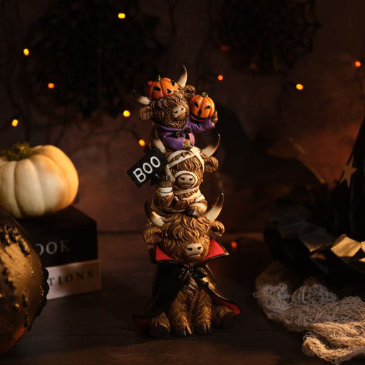 A resin triple stacking highland cow Halloween figurine with three characters — a vampire cow at the bottom in a black and red cape, a mummy cow in the middle holding a BOO sign, and a pumpkin cow at the top in a purple outfit with two jack-o'-lanterns on its horns, styled on a dark surface with Halloween decorations and warm fairy lights in the background