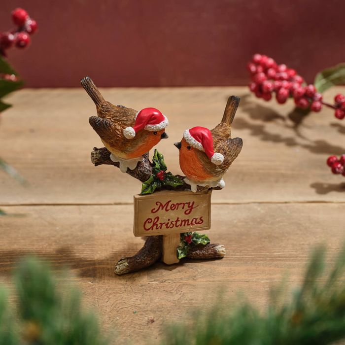 A resin Christmas robin pair figurine with red and white Santa hats, perched on a rustic tree stump with a Merry Christmas sign in red lettering and holly berry detail, on a warm wooden surface with a festive red background and berry sprigs
