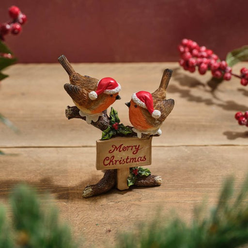 A resin Christmas robin pair figurine with red and white Santa hats, perched on a rustic tree stump with a Merry Christmas sign in red lettering and holly berry detail, on a warm wooden surface with a festive red background and berry sprigs