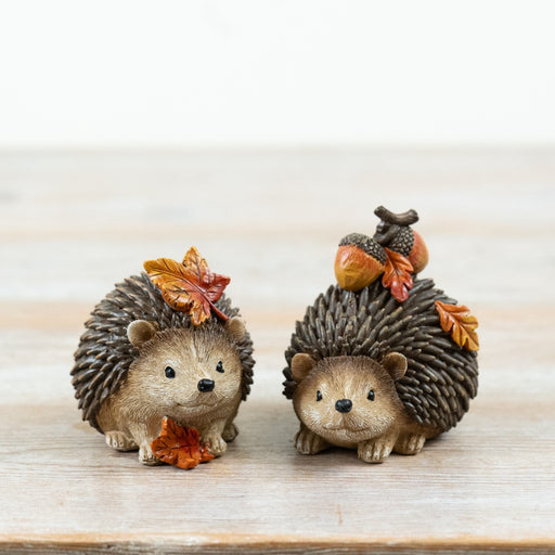 Two autumn hedgehog decorations displayed on a wooden surface, one with autumn leaves and one carrying acorns on his back