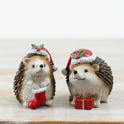 Two Christmas hedgehog decorations in holly-trimmed Santa hats, one holding a red Christmas stocking and one with a small wrapped gift, displayed on a wooden surface