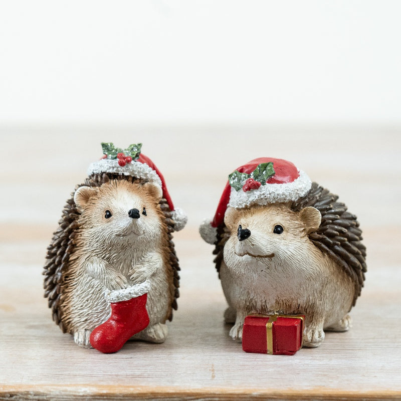 Two Christmas hedgehog decorations in holly-trimmed Santa hats, one holding a red Christmas stocking and one with a small wrapped gift, displayed on a wooden surface