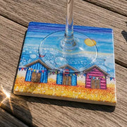 Beach Hut and Bunting ceramic coaster on weathered wooden decking with wine glass showing full coastal scene with beach huts, bunting and bubble-effect sea