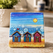 Beach Hut and Bunting ceramic coaster on marble worktop beside plant, showing colourful beach huts with bunting and bubble-effect sea