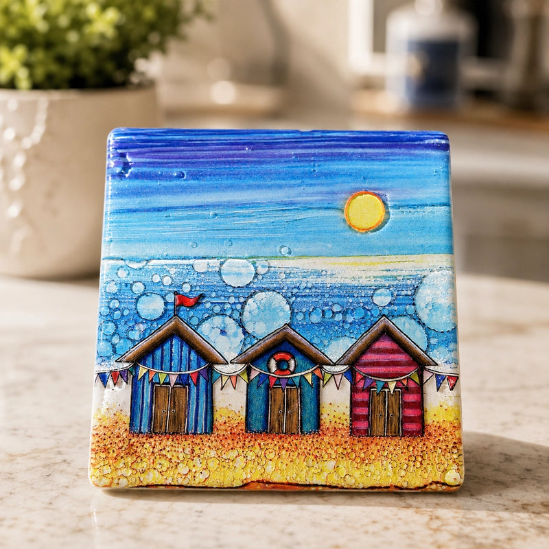 Beach Hut and Bunting ceramic coaster on marble worktop beside plant, showing colourful beach huts with bunting and bubble-effect sea