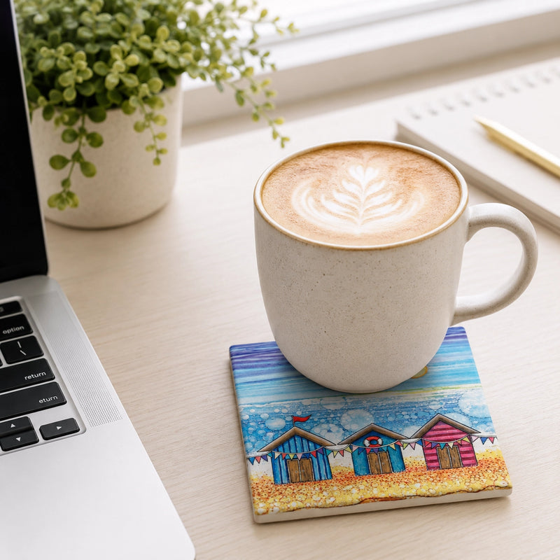 Beach Hut and Bunting ceramic coaster on white desk with latte art coffee mug, laptop, notebook and plant