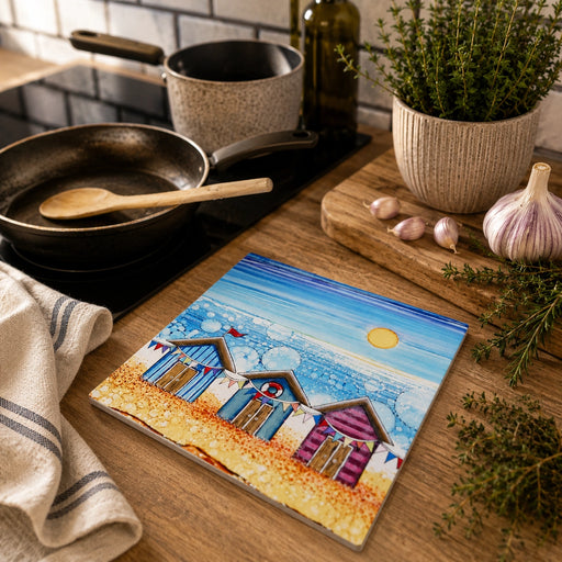 Beach Hut and Bunting ceramic trivet on wooden kitchen worktop beside frying pan and wooden spoon with herb pot in background