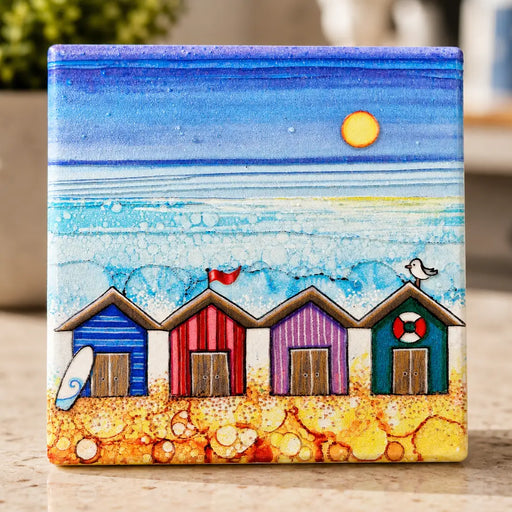 4 Beach Huts ceramic coaster standing upright on kitchen worktop showing full design with four colourful beach huts, surfboard, red flag, seagull and life ring with sparkling textured sea