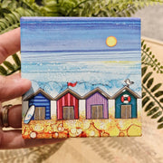 4 Beach Huts ceramic coaster held in hand beside fern plant showing four colourful beach huts with surfboard, red flag, seagull and life ring with sparkling textured sea