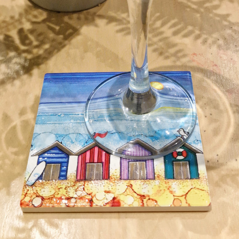 4 Beach Huts ceramic coaster with champagne flute on sandy surface showing full coastal scene with sparkling textured sea