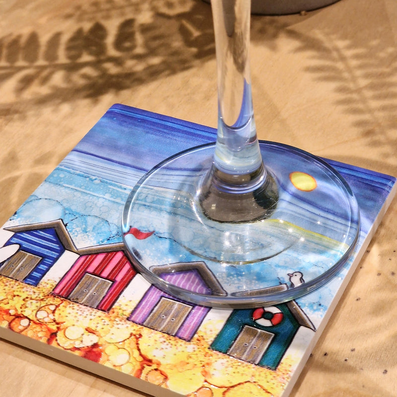 4 Beach Huts ceramic coaster with champagne flute on sandy surface showing full coastal scene with sparkling textured sea