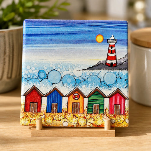 5 Beach Huts and Lighthouse ceramic coaster on wooden easel stand in kitchen setting showing full design with five colourful beach huts, lighthouse and sparkling textured sea