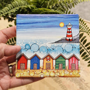 5 Beach Huts and Lighthouse ceramic coaster held in hand beside fern plant showing embossed detail and sparkling textured sea
