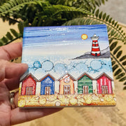5 Beach Huts and Lighthouse ceramic coaster held in hand beside fern plant showing full design with five colourful beach huts and red and white striped lighthouse