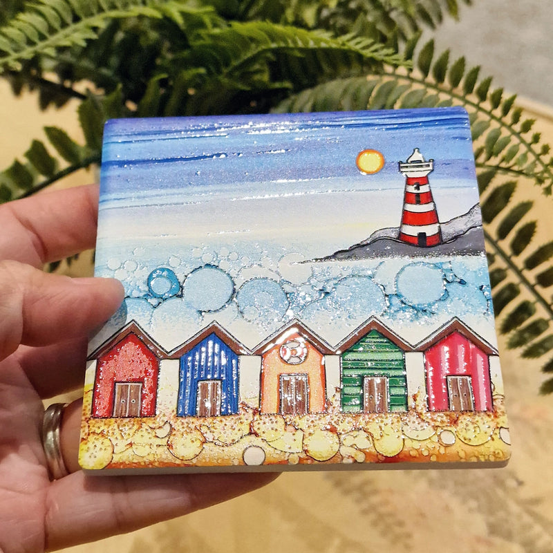 5 Beach Huts and Lighthouse ceramic coaster held in hand beside fern plant showing full design with five colourful beach huts and red and white striped lighthouse