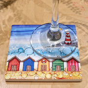 5 Beach Huts and Lighthouse ceramic coaster on sandy surface with champagne flute showing full coastal scene