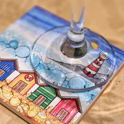 5 Beach Huts and Lighthouse ceramic coaster on sandy surface with champagne flute, close-up showing embossed beach hut and lighthouse detail